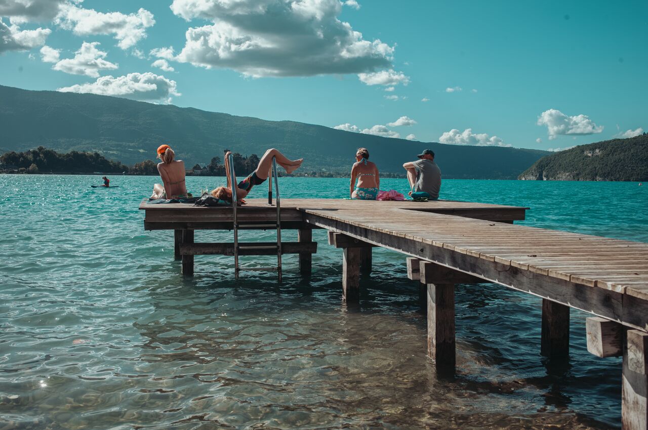 Four people relax on a wooden dock at Lake Annecy, sitting and sunbathing beside clear water.