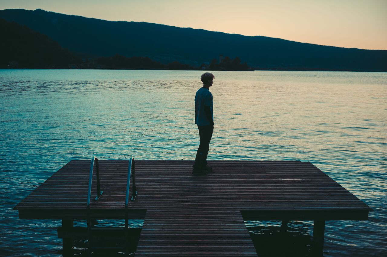 Person stands on a wooden dock at Lake Annecy, facing the water at dusk, with mountains in the background.
