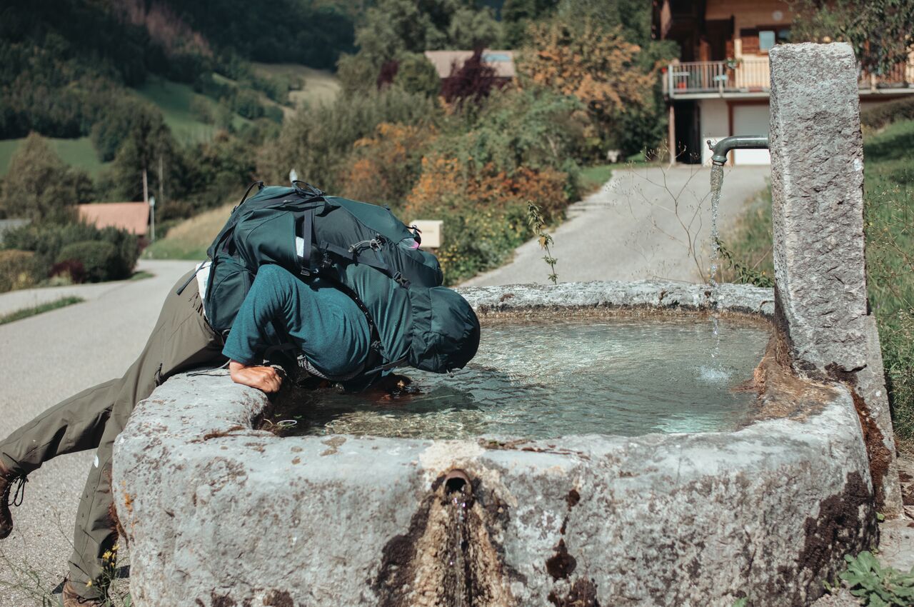 A hiker with a large backpack bends over a stone fountain, dunking his head in the water to cool off.