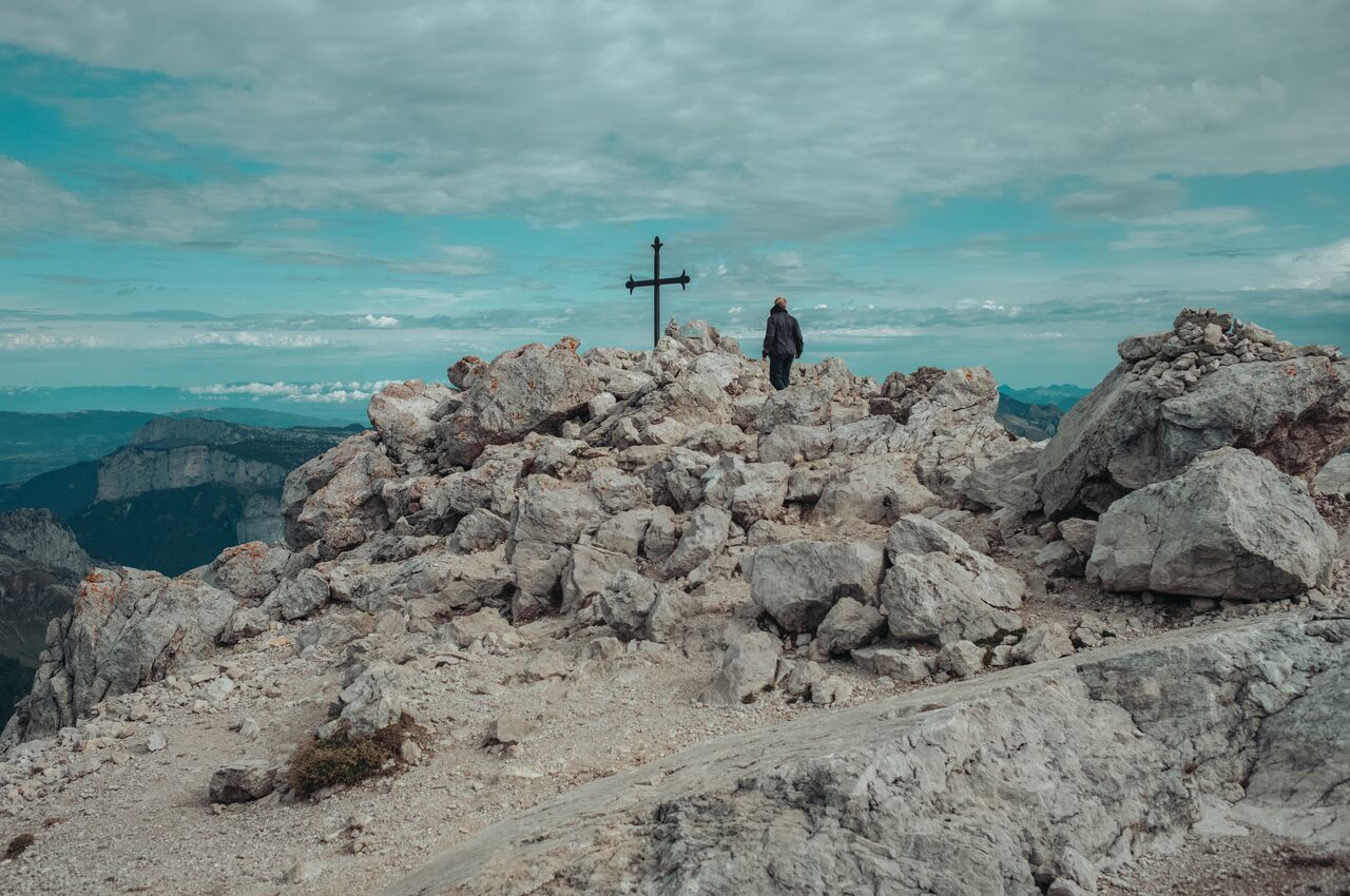 A hiker stands beside a metal cross on a rocky summit, surrounded by large boulders, looking out over distant mountains under a cloudy sky.