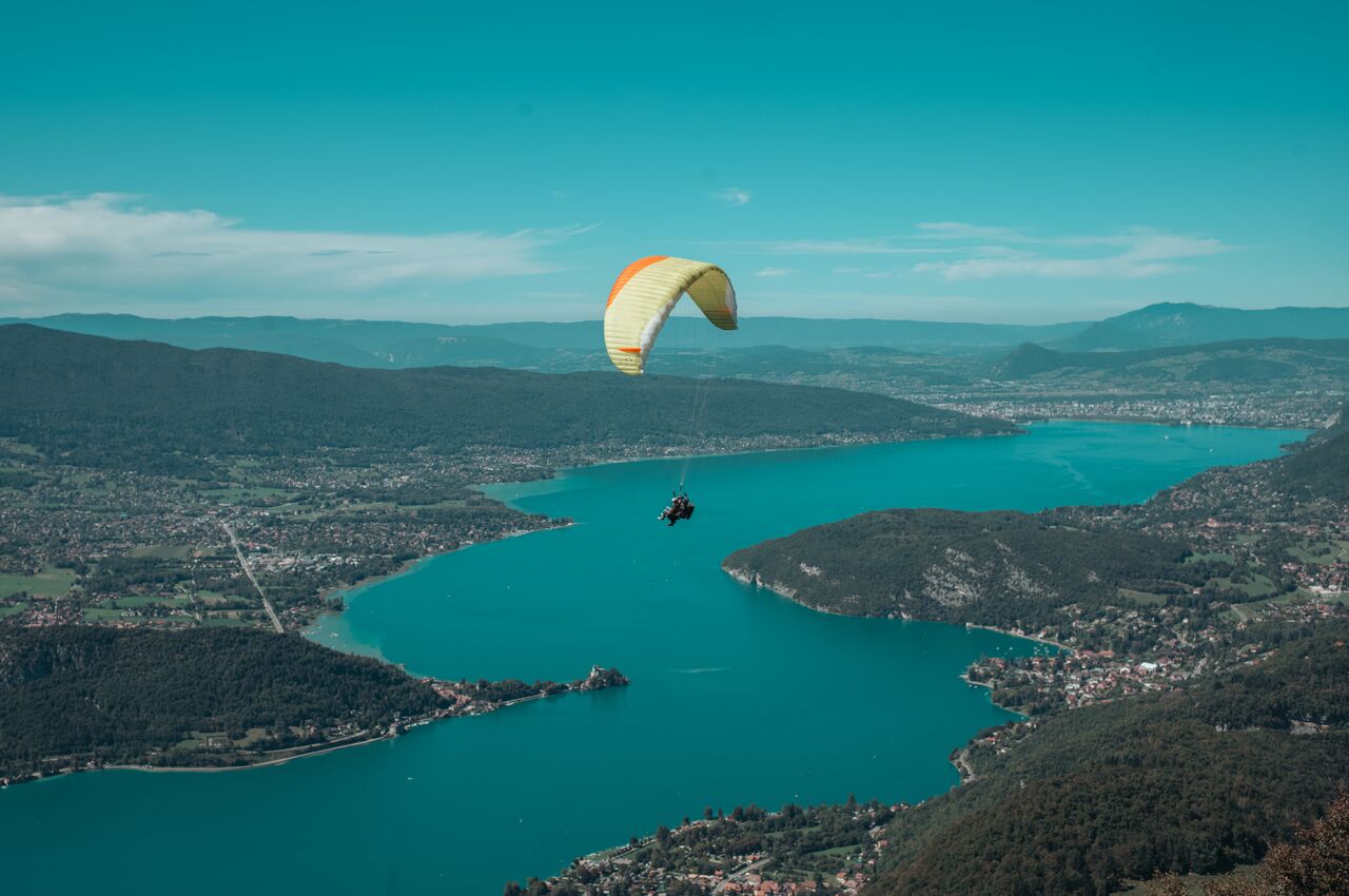 A paraglider glides high above Lake Annecy, the blue water curving between green hills with small towns scattered along the shore below.