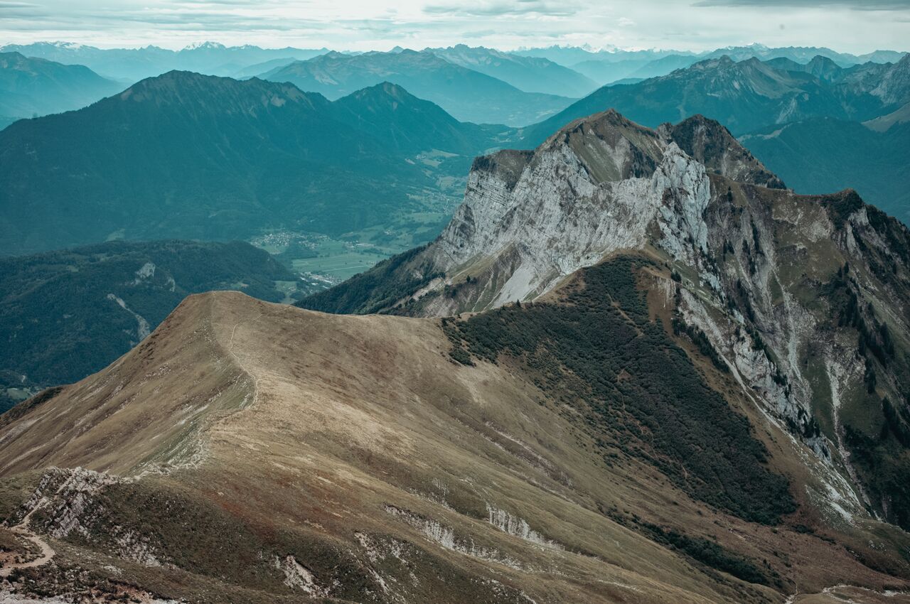 A narrow hiking trail runs along a grassy ridge toward rocky peaks, then drops into the forest.
