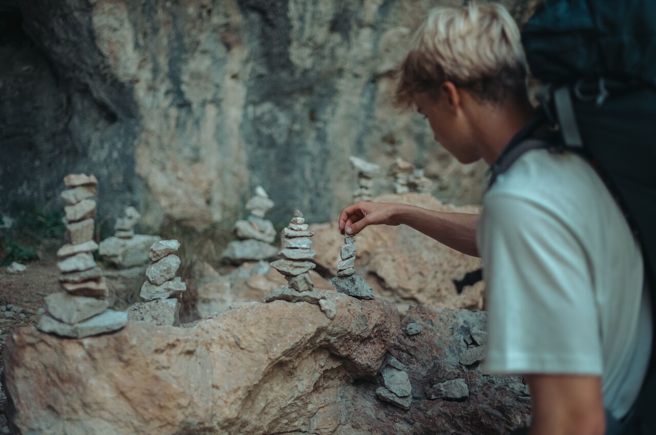 A hiker with a backpack adds a small rock to a stacked cairn on a rocky ledge.