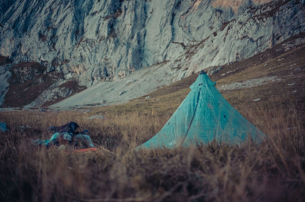 A green pyramid tent stands on a grassy meadow below a steep rocky mountainside, with camping gear scattered nearby.