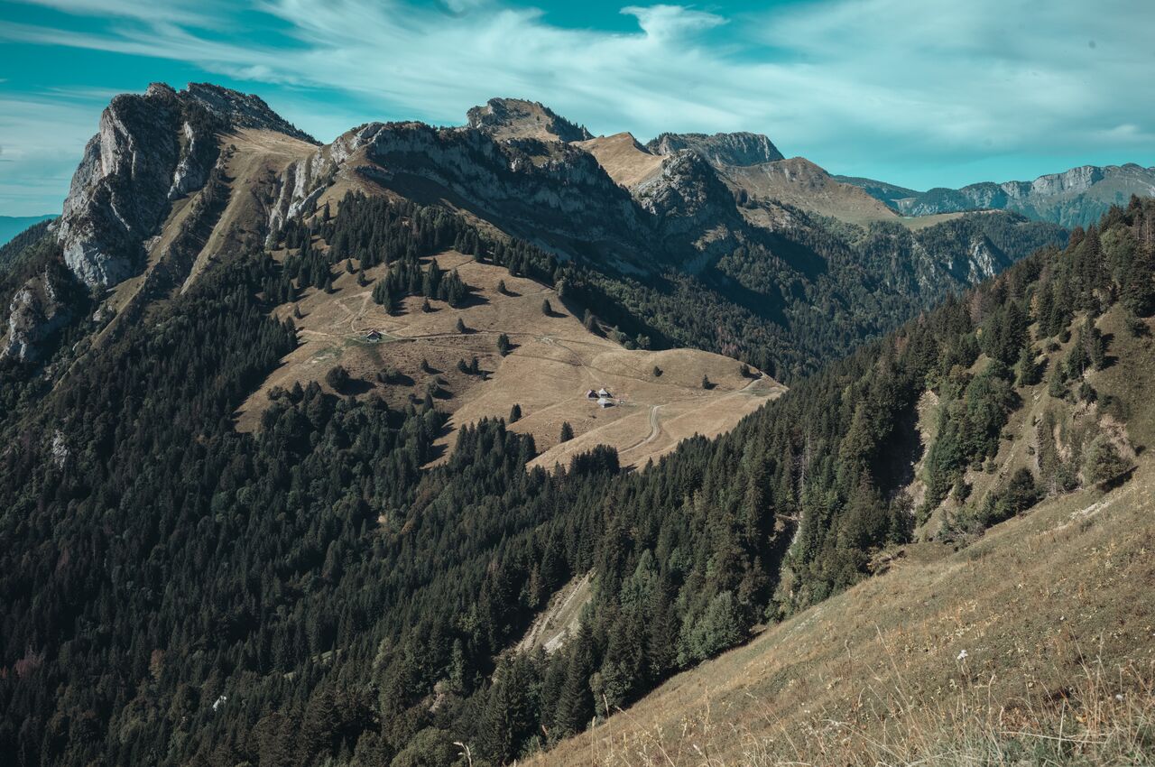 View from the trail near La Tournette: mountain ridges and forested slopes, with a small chalet in a grassy clearing below.