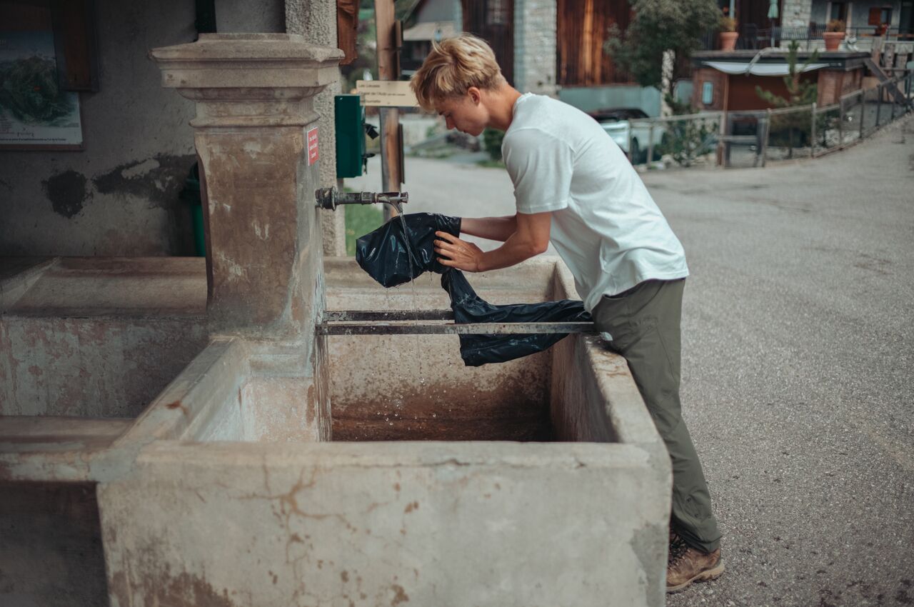 A person rinses muddy rain pants under a tap at a stone fountain.