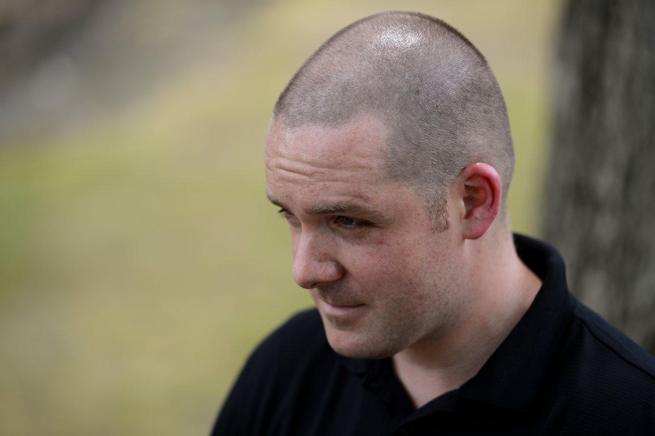 A man in a black shirt looks to the side with a neutral expression at an outdoor gathering.