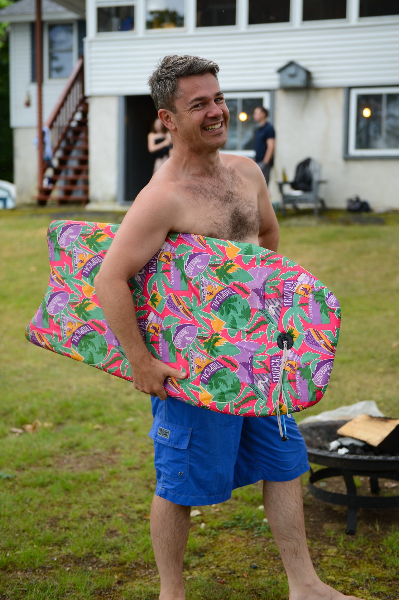 A smiling man in blue shorts holds a colorful boogie board outdoors near a house with people in the background.