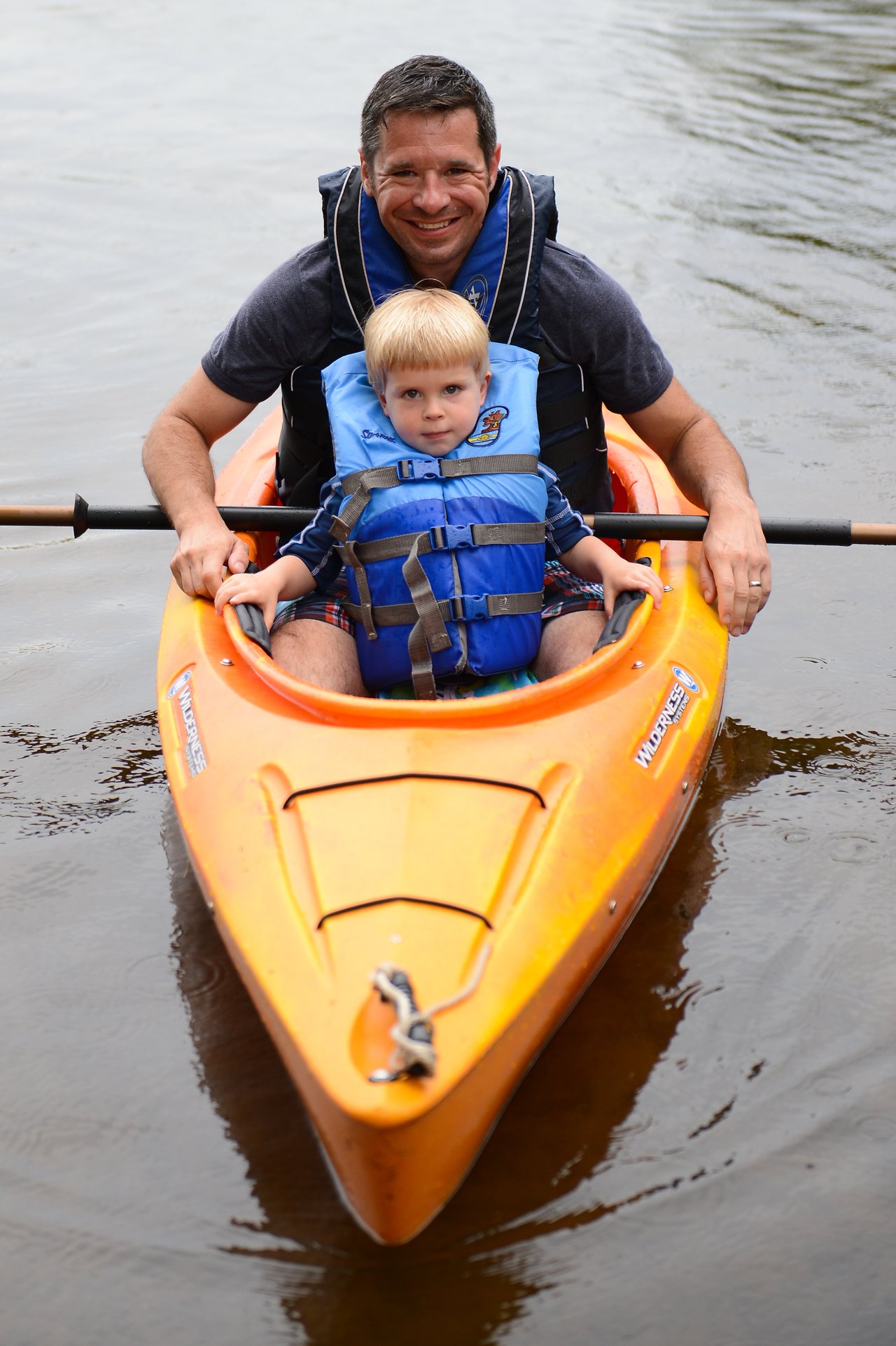 A man and a young child wearing life jackets sit in a bright orange kayak on calm water.