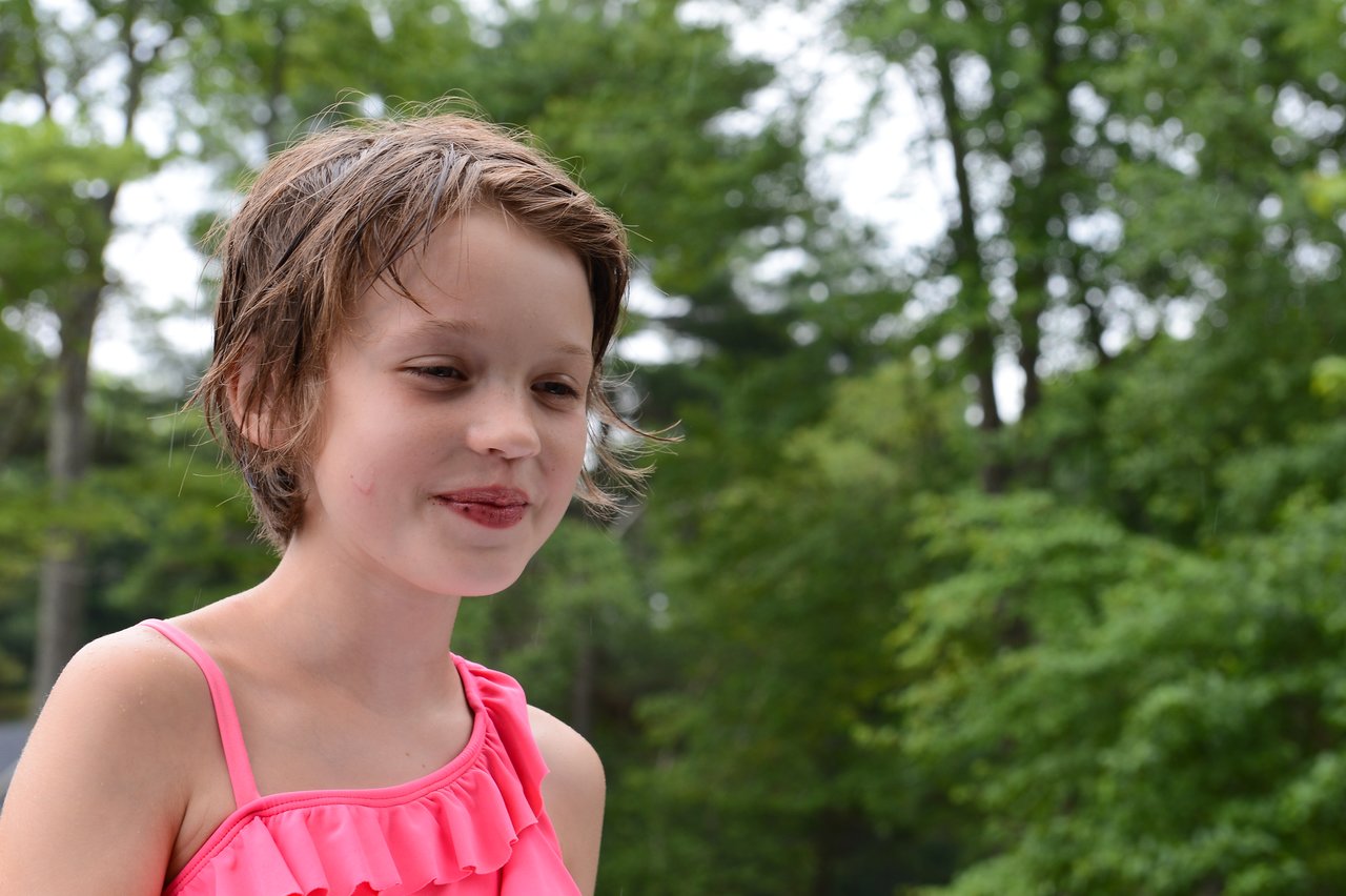 A young girl in a pink swimsuit smiles with wet hair, standing outdoors with trees in the background.