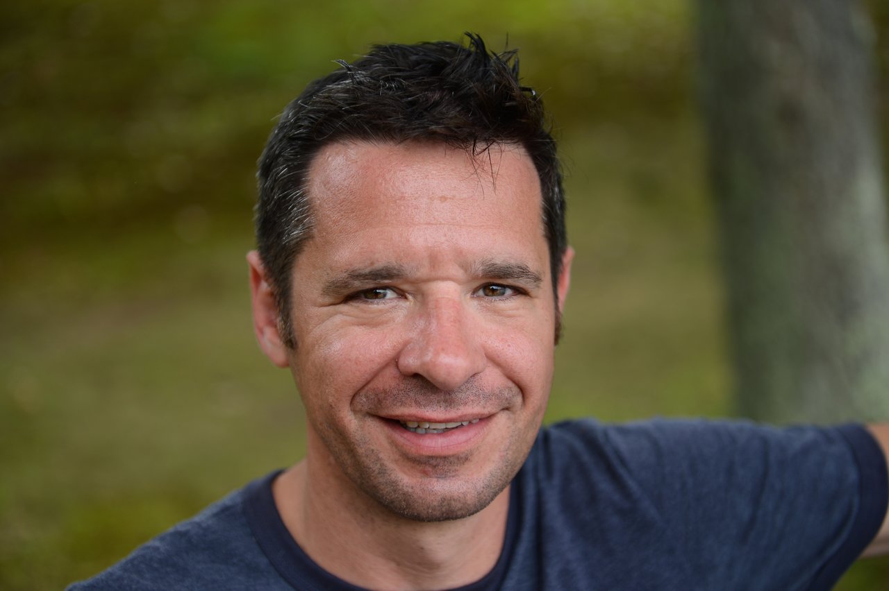 A man in a navy blue shirt smiles while sitting outdoors at a gathering.