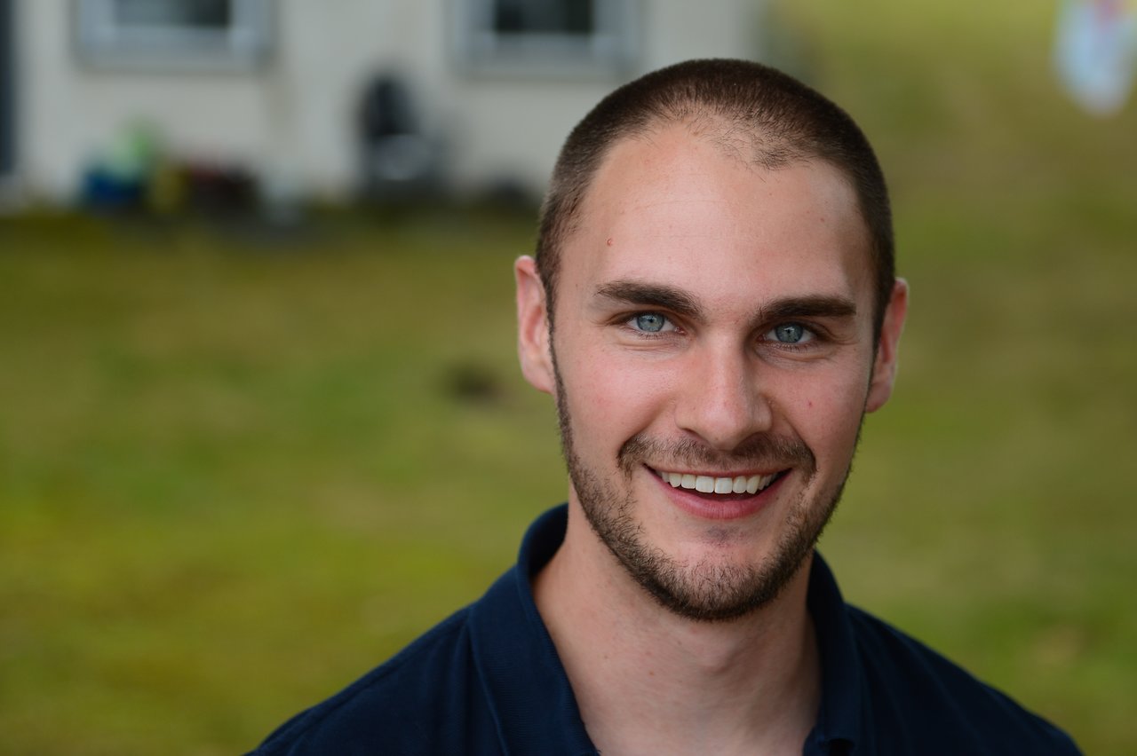 A man with short hair and a beard smiles at the camera outdoors during a gathering.