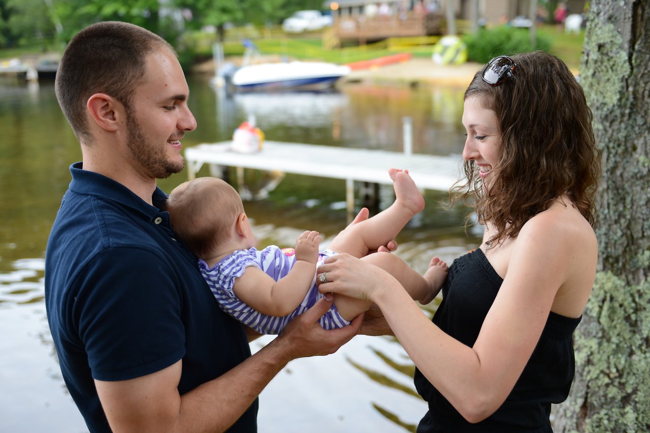 A man holds a baby while a woman playfully touches the baby's feet near a lakeside dock.