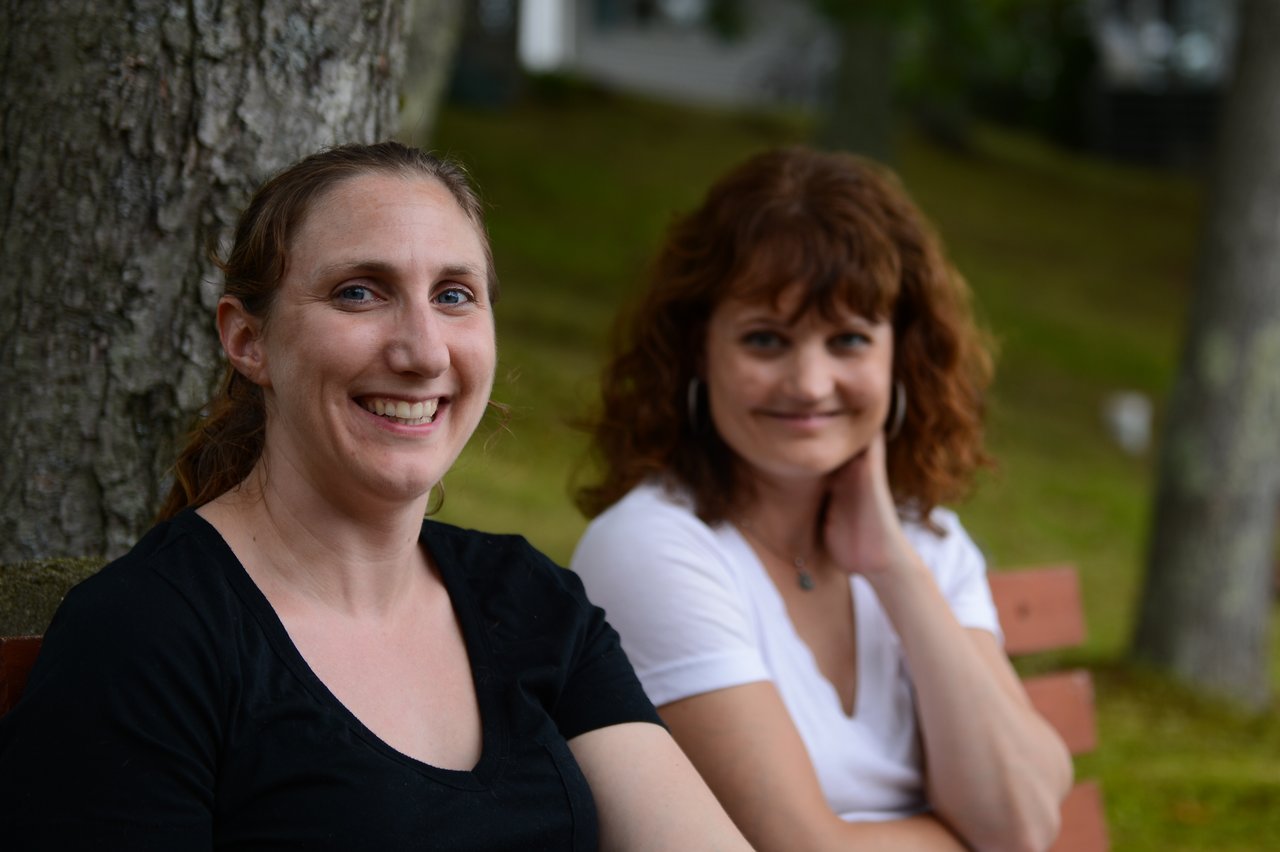Two women sitting on a bench outdoors, one smiling at the camera while the other looks slightly away.