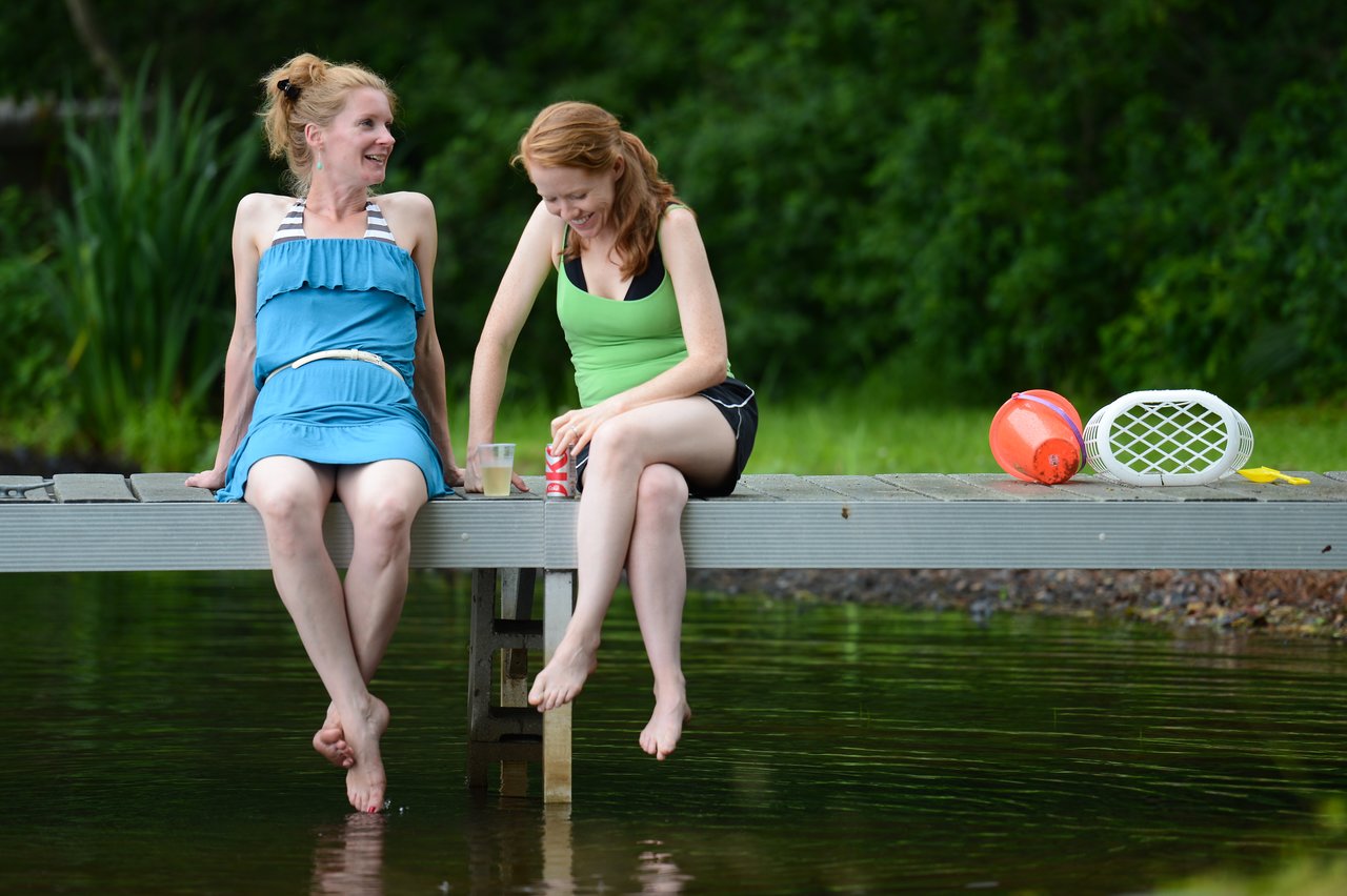 Two people sit on a dock, smiling and dipping their feet in the water, with drinks and toys nearby.