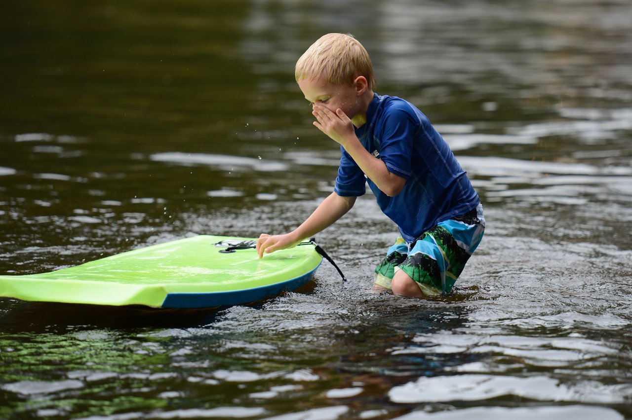 A young child in swimwear kneels in shallow water, touching a green boogie board while wiping their face.