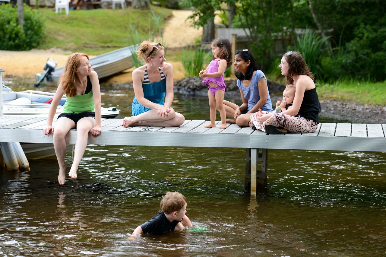 A group of women and children sit on a dock, talking, while a boy swims in the water below.