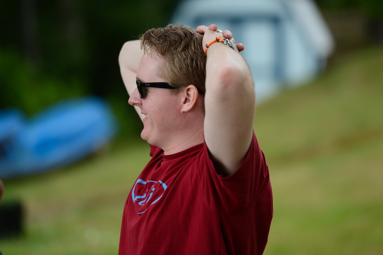A man in a red shirt and sunglasses smiles with his hands resting on his head outdoors.