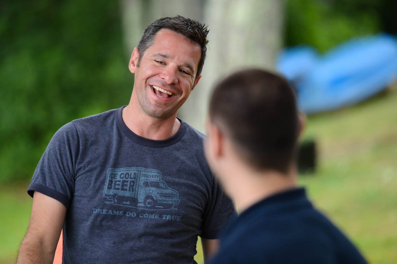 A man in a gray t-shirt smiles and talks with another person at an outdoor gathering.