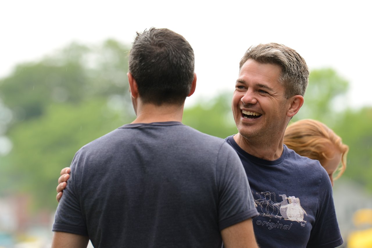 A man smiles while greeting another person with a hand on their shoulder at an outdoor gathering.