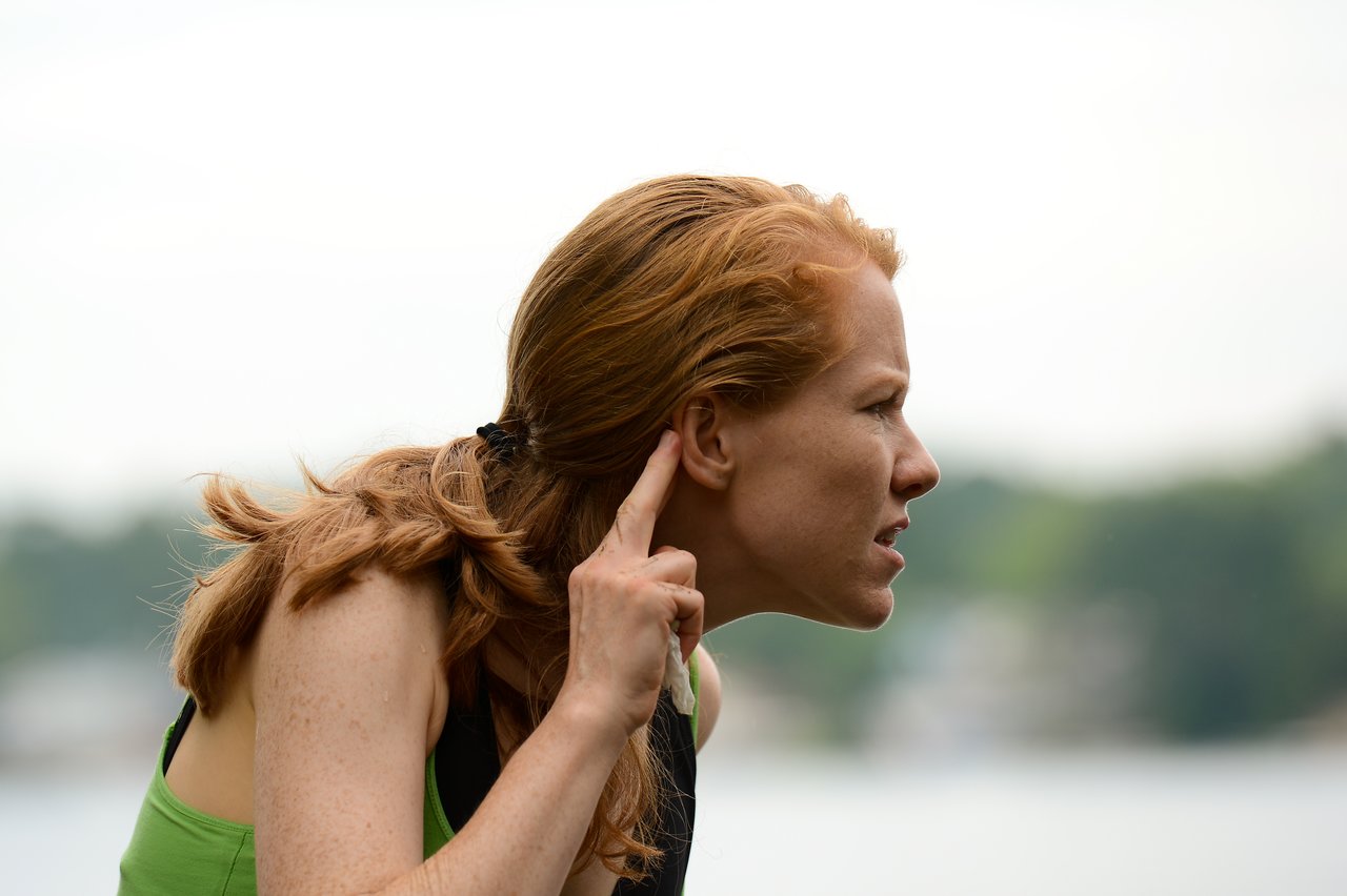 A woman in a green top points to her ear, appearing to listen closely during an outdoor gathering.