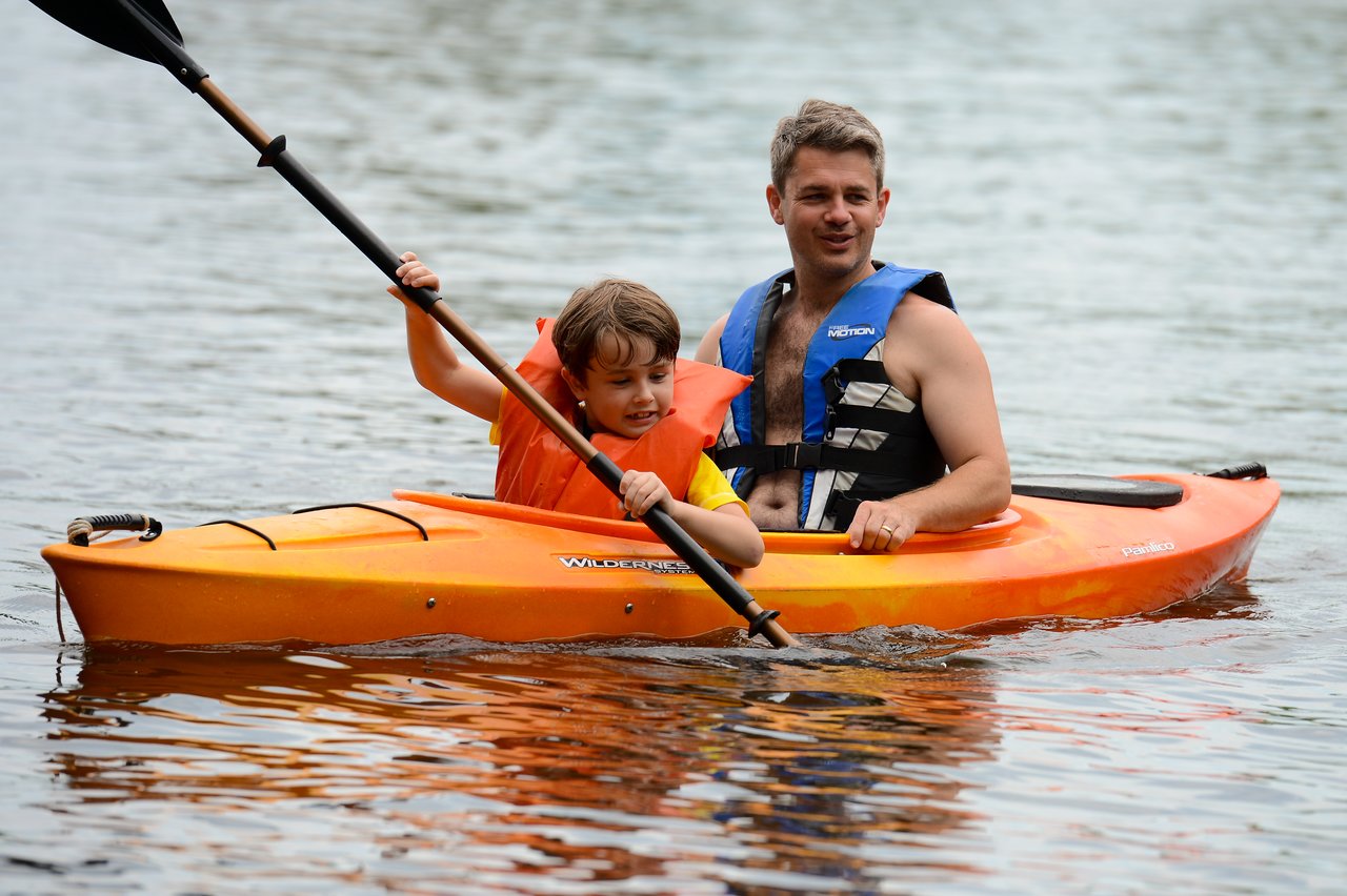 A child and an adult wearing life jackets paddle together in an orange kayak on the water.