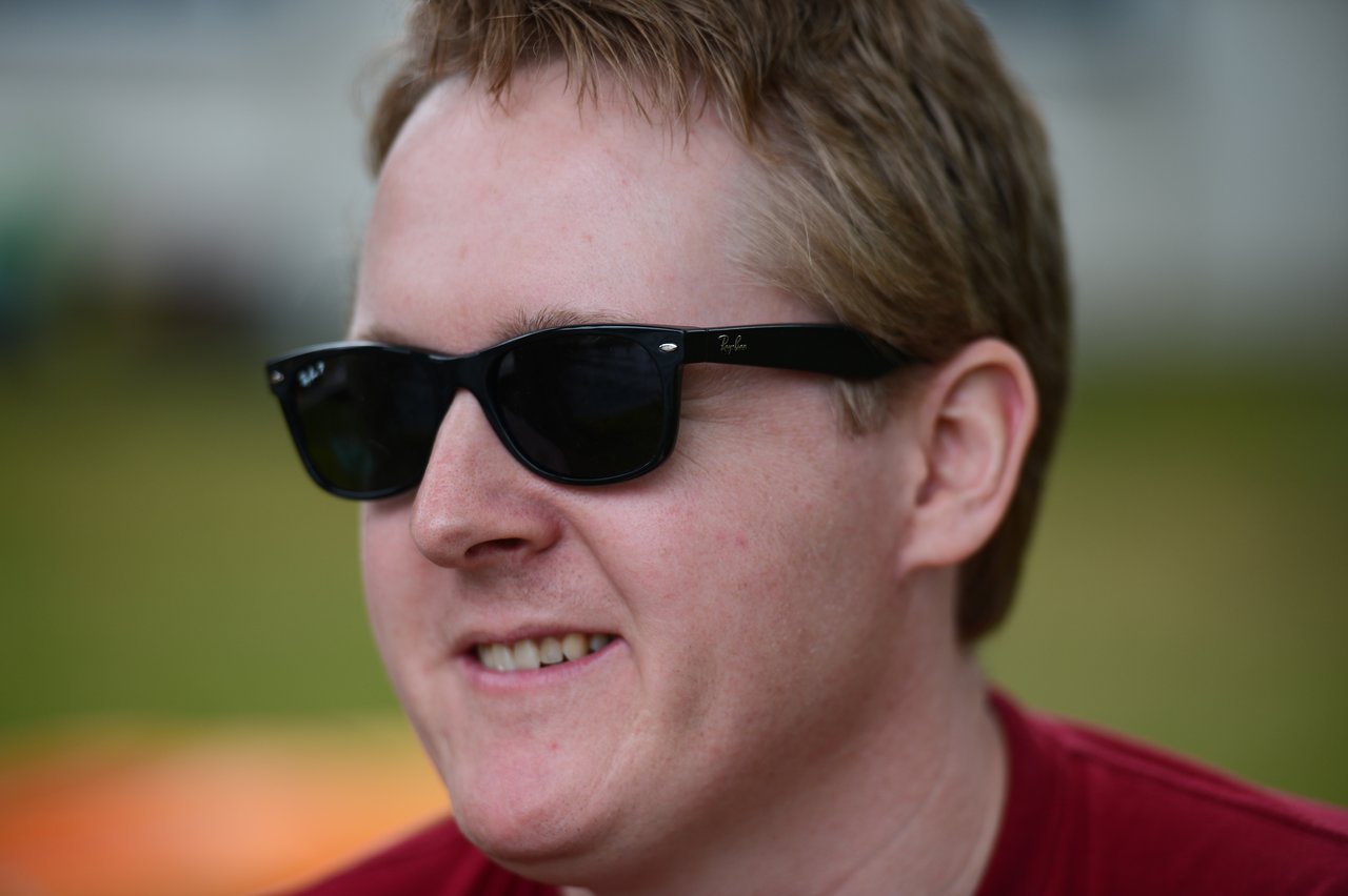 A person wearing sunglasses and a red shirt smiles while outdoors at a gathering.