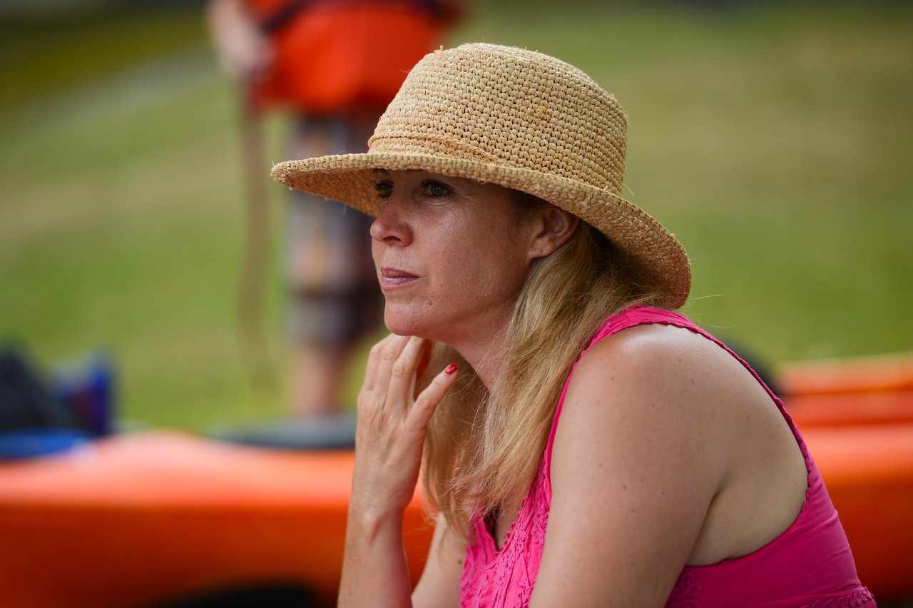 A woman in a straw hat and pink top sits outdoors, looking thoughtful.