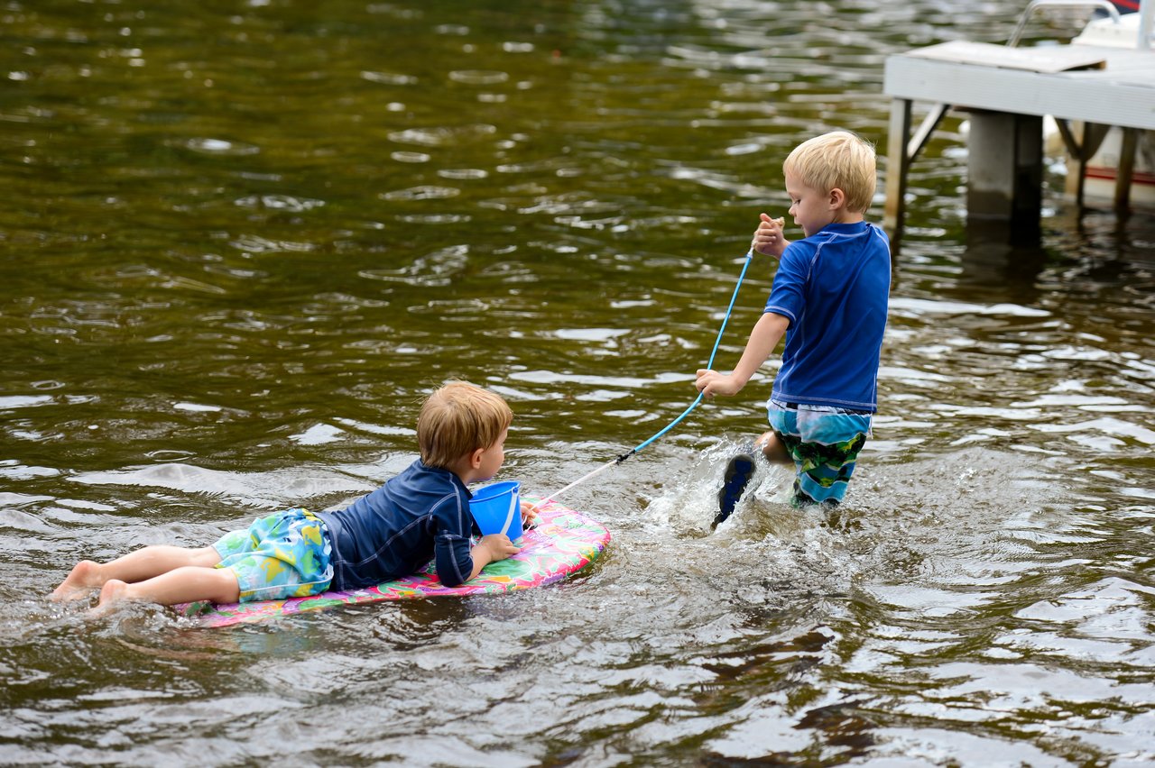 A child pulls another child on a float through shallow water, both wearing swim clothes and playing near a dock.