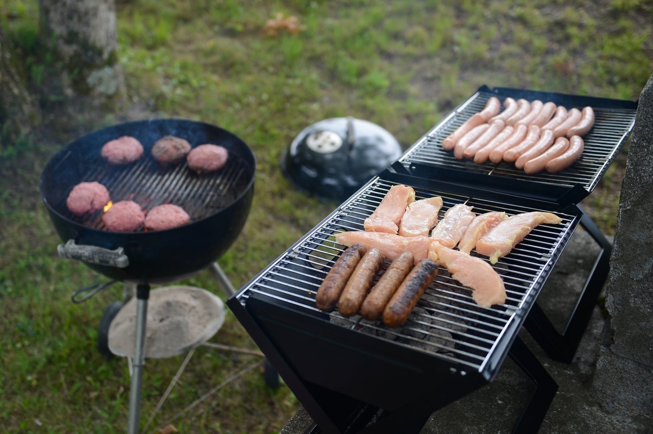 Multiple grills with burgers, sausages, and chicken cooking outdoors on a grassy area.