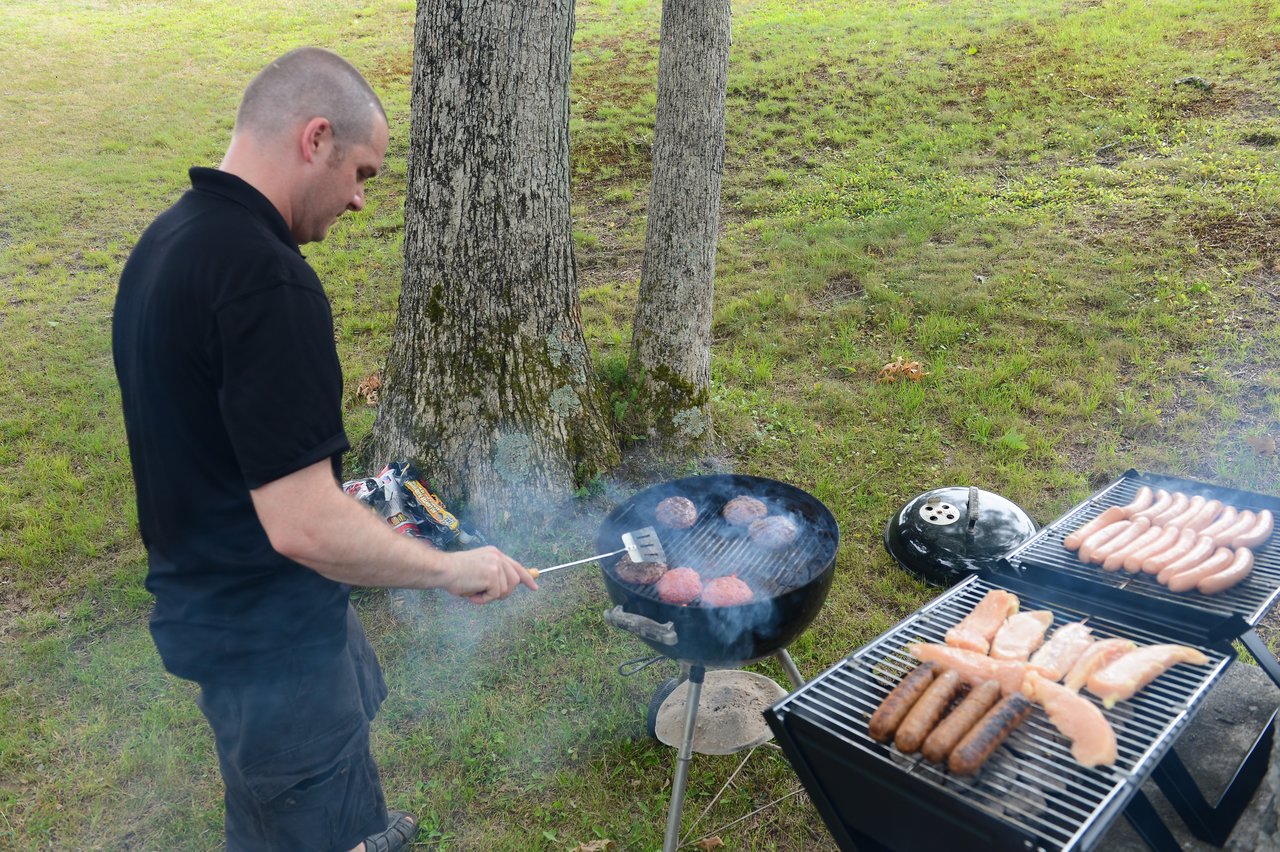 A man grills burgers, sausages, and chicken on two barbecues at an outdoor gathering.