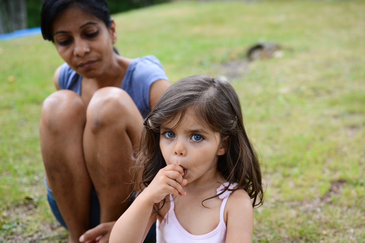 A young girl in a pink top sucks her thumb while a woman sits nearby on the grass.
