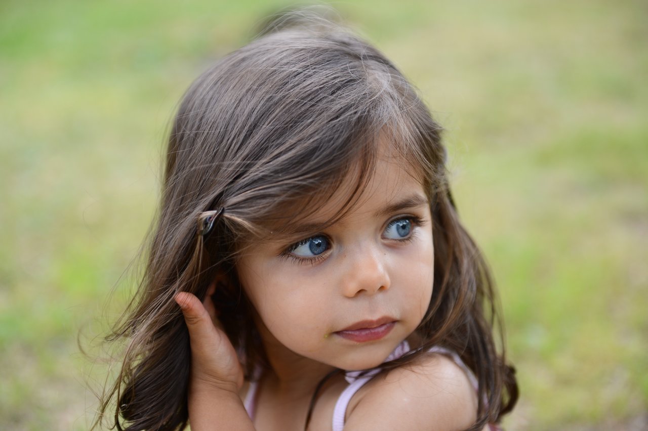 A young girl with long brown hair looks to the side while touching her hair outdoors.