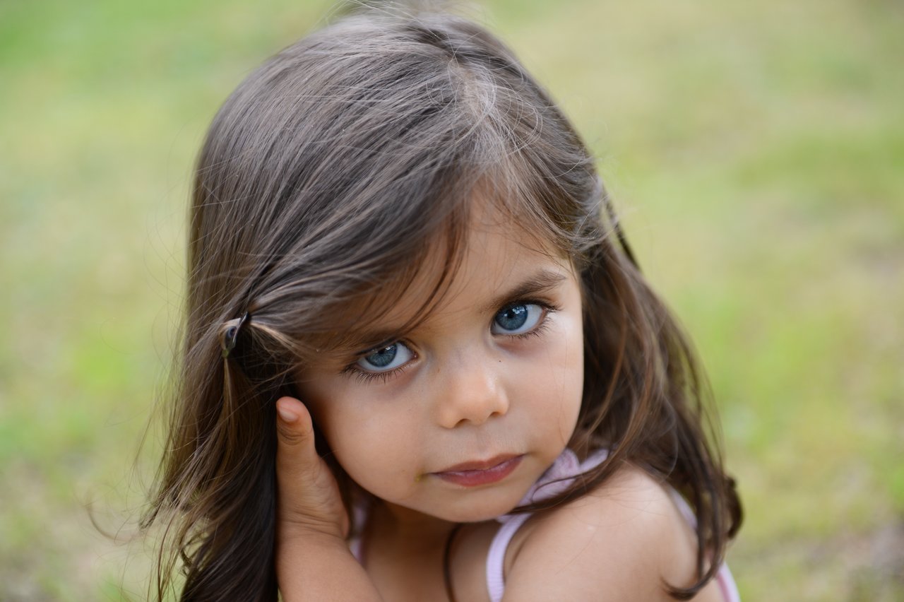 A young girl with long brown hair looks at the camera while touching her ear, standing outdoors on grass.