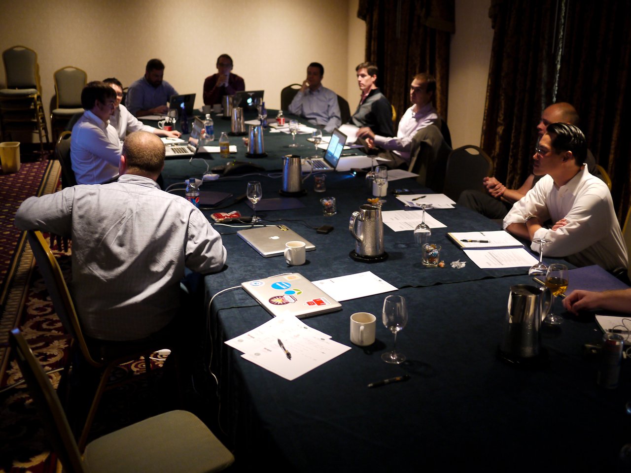 A group of people in a meeting room sit around a table with laptops, papers, and drinks.