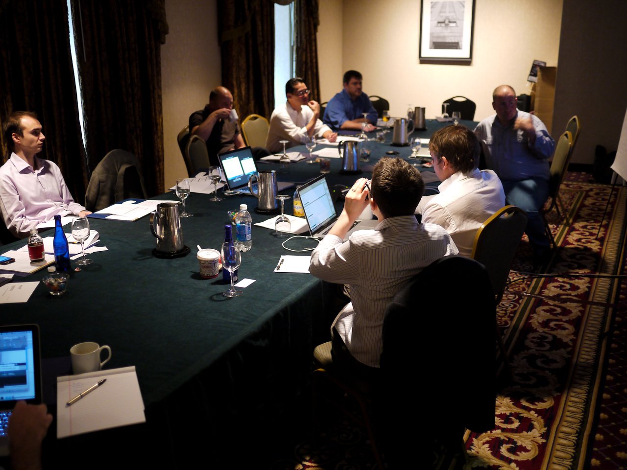 A group of people in a conference room engaged in a discussion, with laptops and notes on the table.