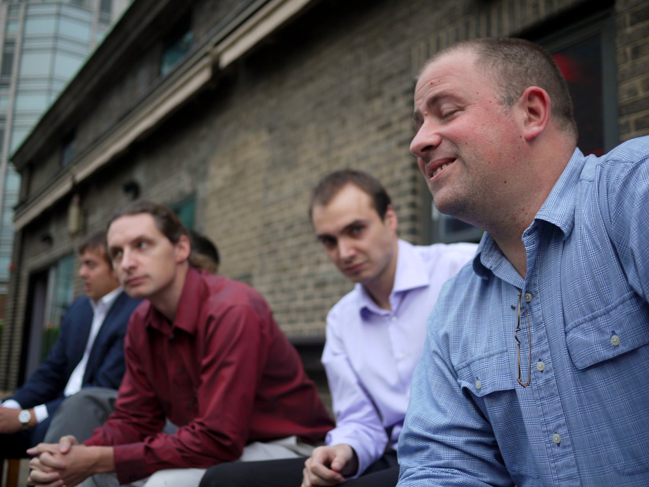 Four men sit outside near a brick building, with one in a blue shirt smiling and speaking.