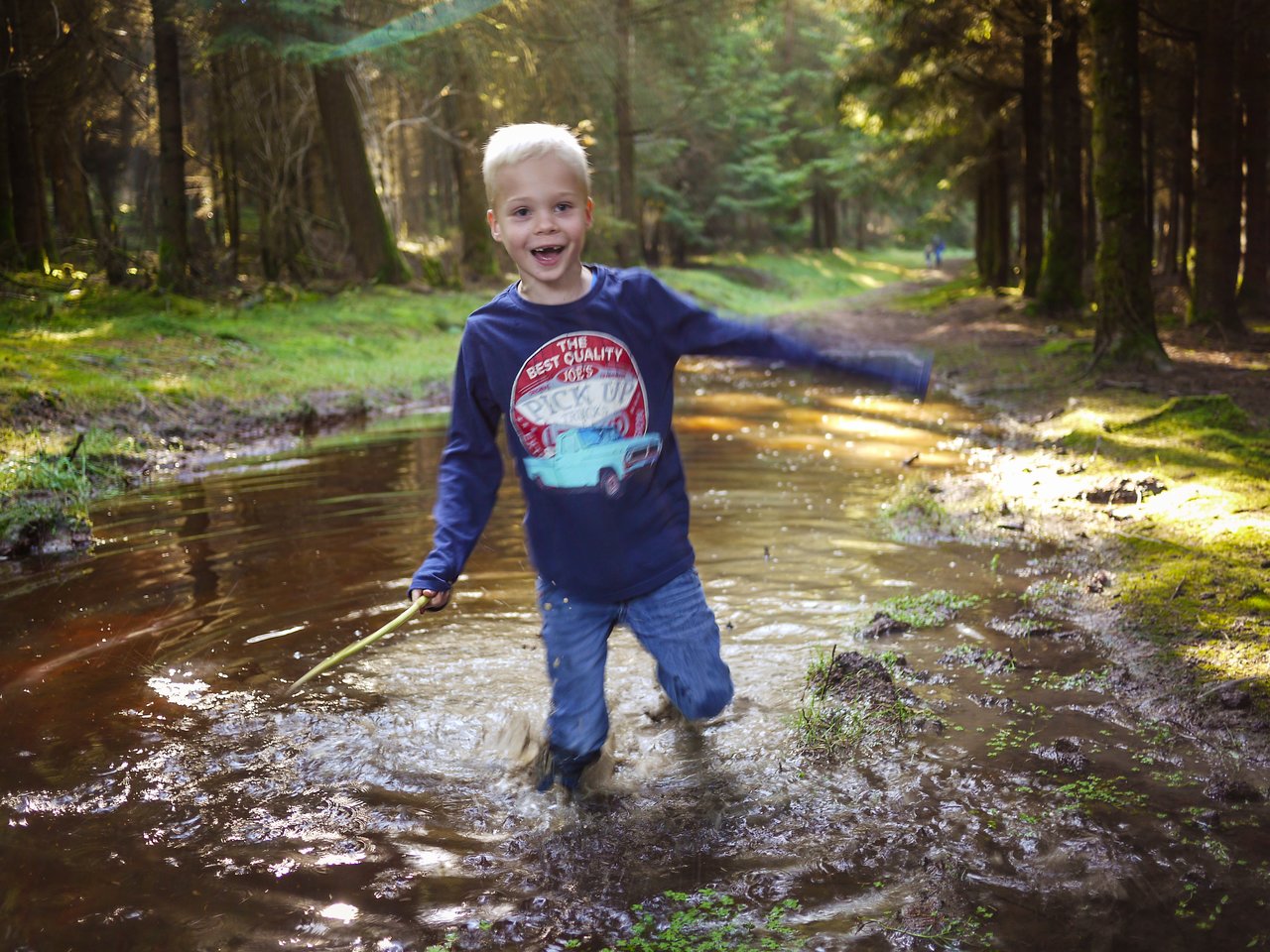 A young boy with a stick splashes through a muddy puddle in a forest, smiling and playing.