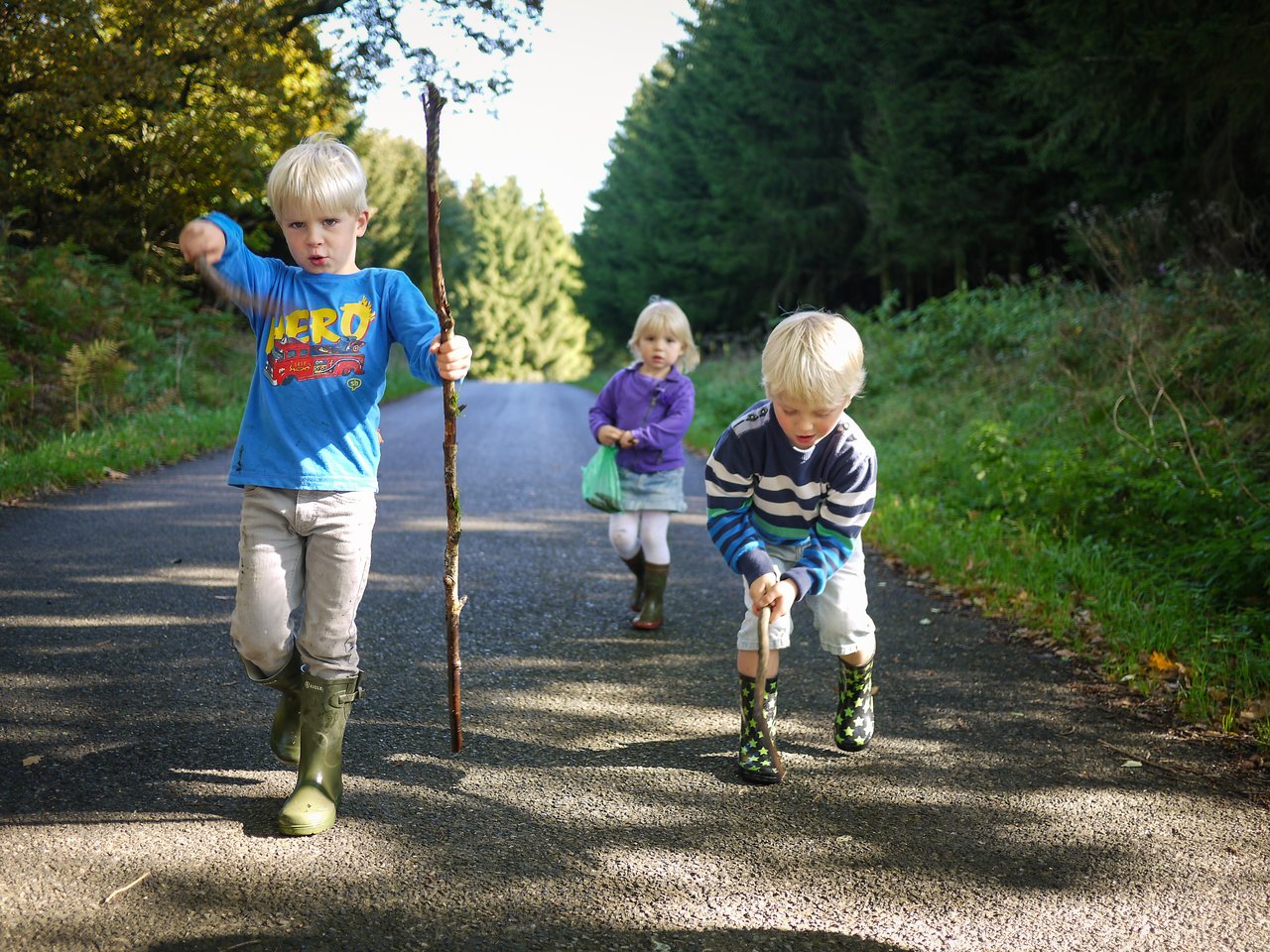 Three young children walk on a paved road, two holding sticks, while one follows carrying a small bag.