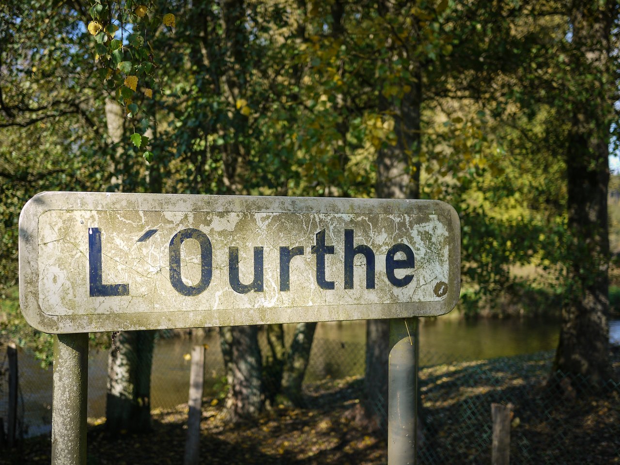A weathered sign reading "L'Ourthe" stands in front of trees and a river in Lavacherie.