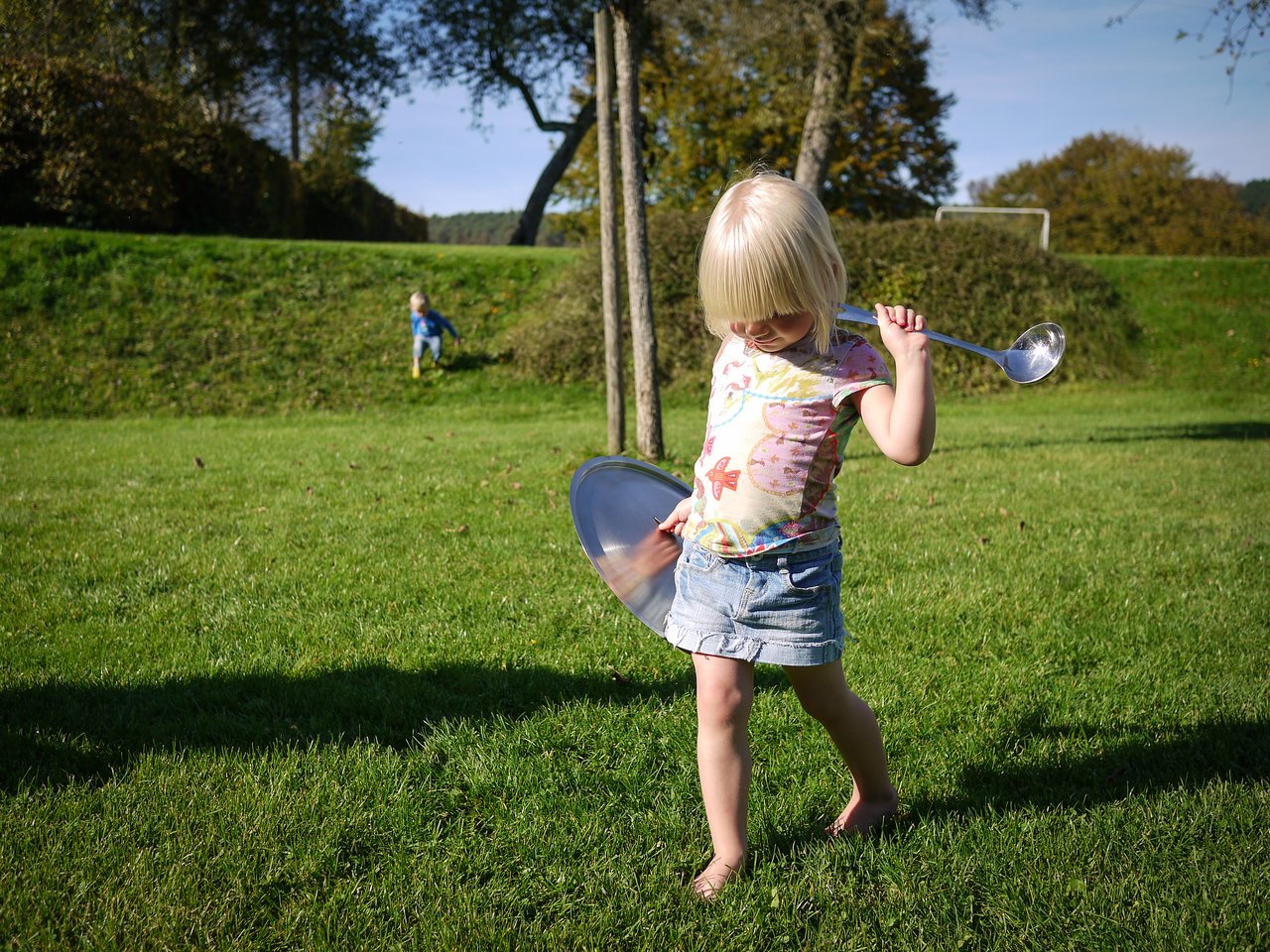 A young child walks barefoot on grass, holding a large spoon and metal lid, with another child in the background.