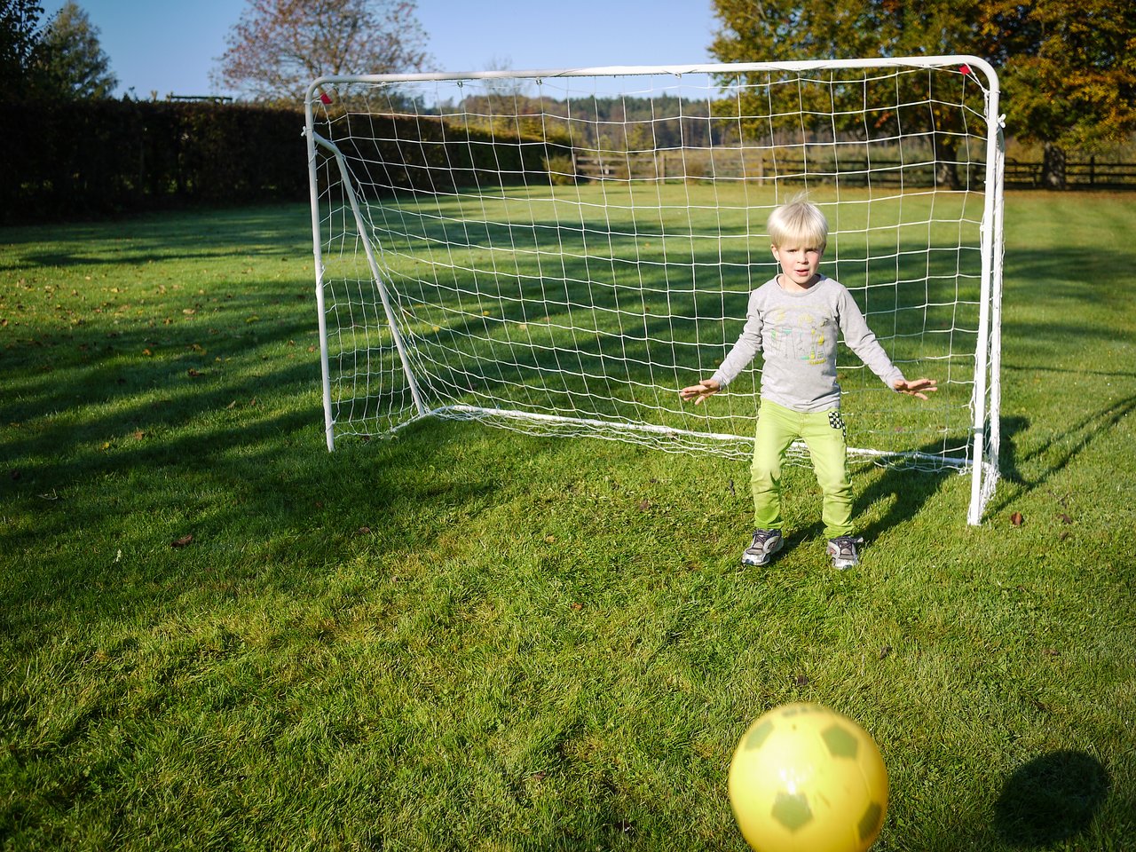 A child stands in front of a small soccer goal, preparing to block a yellow ball rolling toward them.