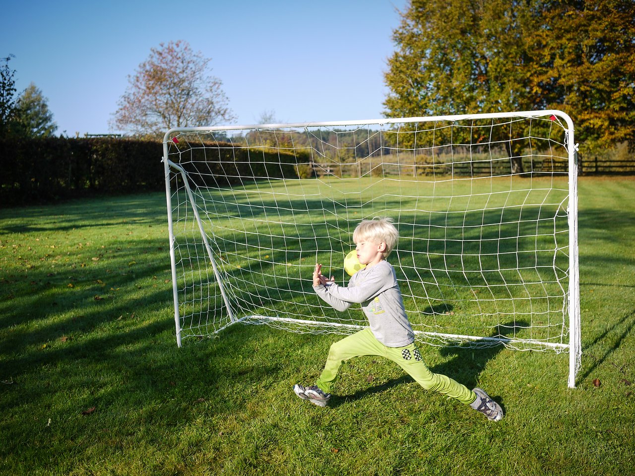A child in a gray sweater and green pants tries to block a soccer ball in front of a goal.