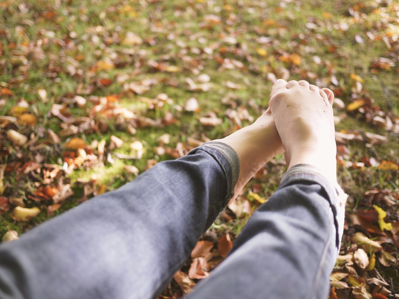 A person sits on grass with fallen leaves, wearing jeans, holding their bare feet with both hands.