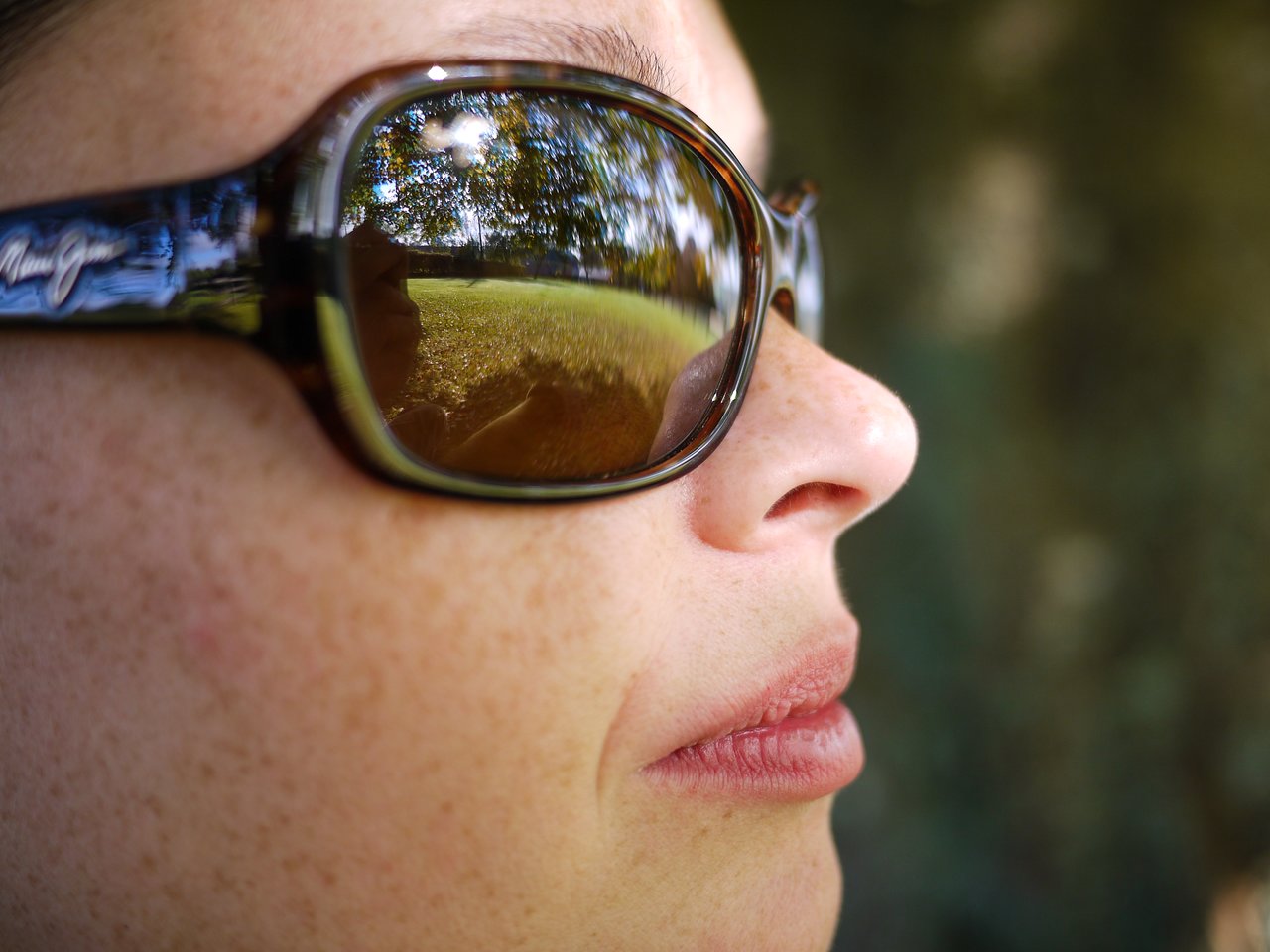 Close-up of a person wearing sunglasses, reflecting a grassy field with trees and sunlight in the lenses.