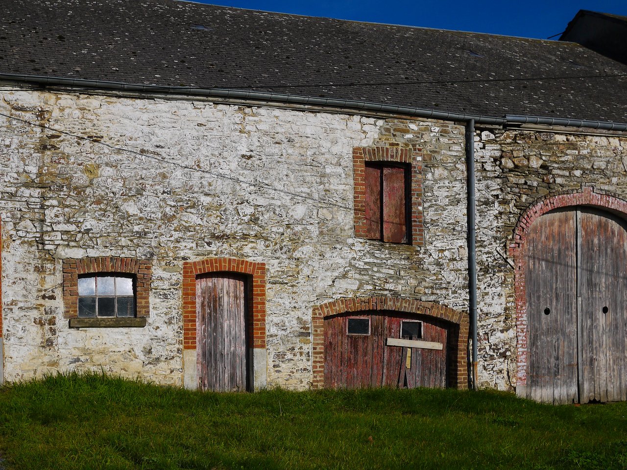 An old stone barn with wooden doors and windows, surrounded by grass under a clear blue sky.