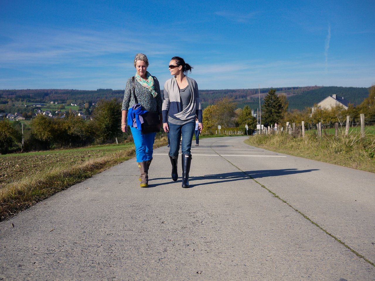 Two women walk along a paved rural road, talking and enjoying the sunny weather.