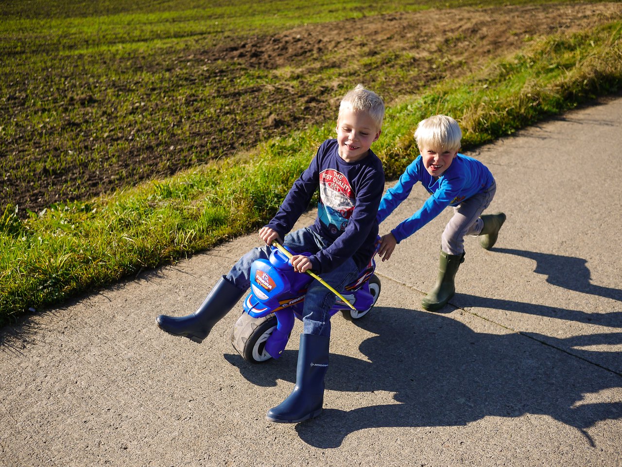 A child rides a small toy motorcycle while another child pushes from behind on a paved path.