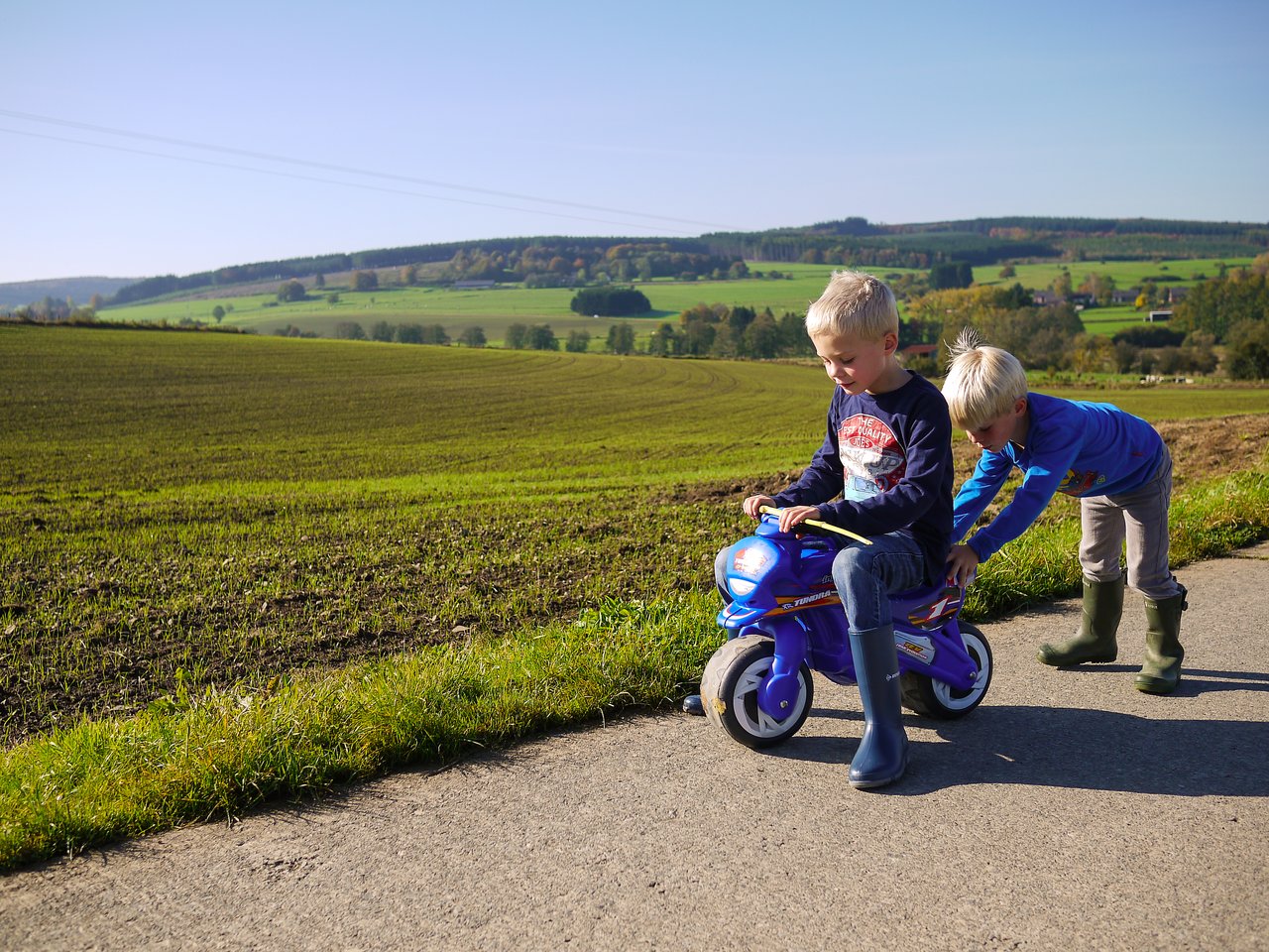 A child rides a small toy motorcycle while another child pushes from behind on a rural path.