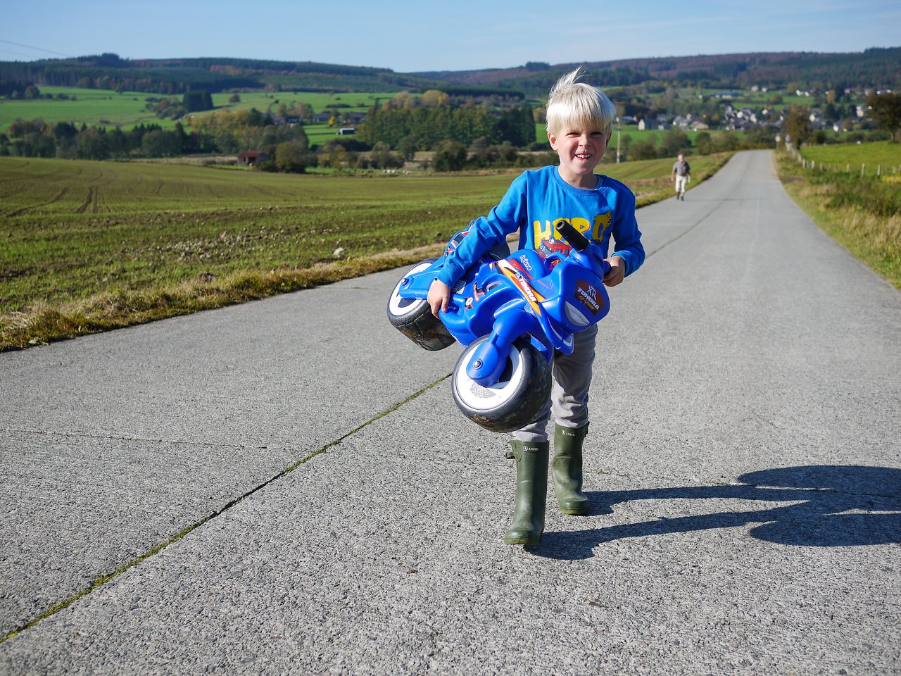 A smiling child in boots carries a blue toy motorcycle while walking on a rural road.
